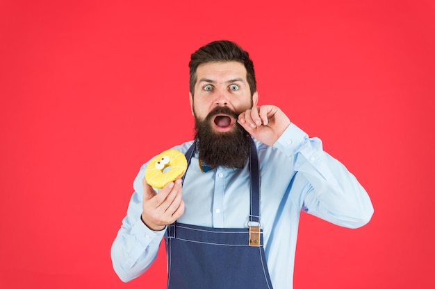 カフェとパン屋のコンセプトパン屋からの甘いドーナツ料理エプロンの男のひげを生やしたパン屋はかわいいデザートを保持しますダイエットを忘れるチートミールヒップスターのひげを生やしたパン屋は赤い背景に艶をかけられたドーナツを保持します