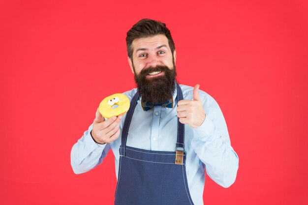 Cafe and bakery concept Sweet donut from baker Man bearded baker in cooking apron hold cute dessert Ways to reduce hunger and appetite Hipster bearded baker hold glazed donut on red background