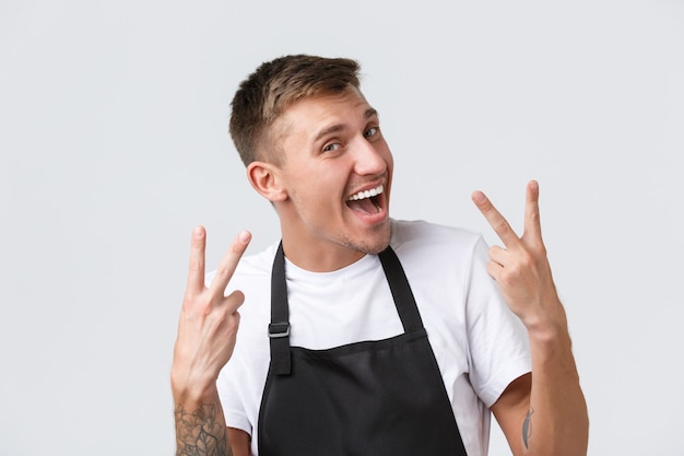 Cafe and restaurants, coffee shop owners and retail concept. Cheerful handsome caucasian guy waiter, barista in black apron showing victory sign and smiling upbeat, white background