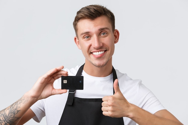 Cafe and restaurants, coffee shop owners and retail concept. Cheerful handsome salesman, store clerk in black apron showing thumbs-up in approval, recommend paying for credit card