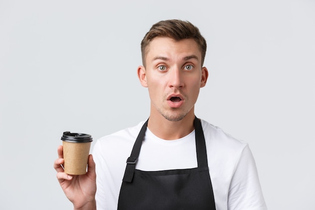 Cafe and restaurants coffee shop owners and retail concept startled and confused barista waiter in black apron holding paper cup with takeaway drink white wall