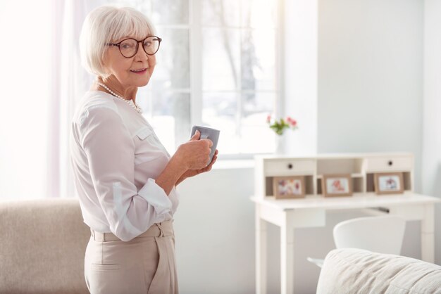 Caffeine lover. Pleasant pretty elderly woman standing half-turned in the living room and posing while holding a cup of coffee