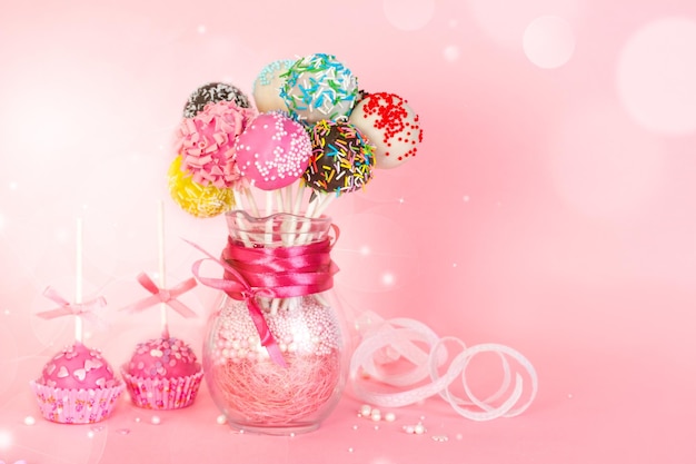 写真 紙の形にピンクのアイシングと装飾が施されたケーキポップとピンクの背景にガラスの花瓶のカラフルなケーキポップ