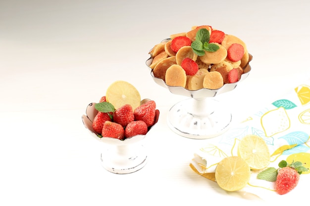 흰색 바탕에 민트 잎과 레몬 조각으로 장식된 작은 팬케이크 시리얼과 딸기가 있는 케이크 스탠드. 트렌디한 음식. 미니 시리얼 팬케이크. 복사 공간이 있는 가로