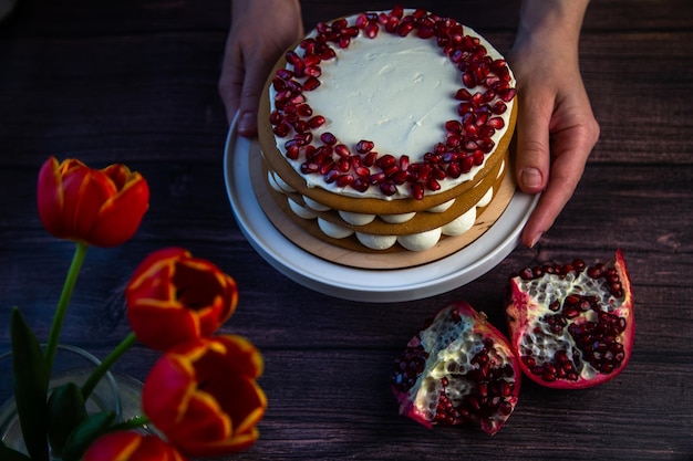 The cake stands on a white stand decorated with cream and pomegranate on top it is held by the hands...