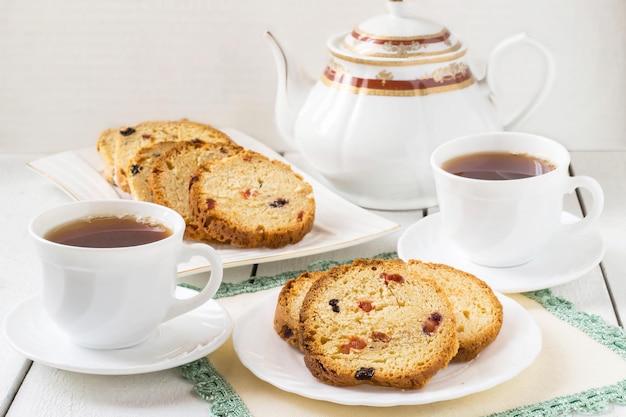 Cake with raisins on a plate, white cup of tea and teapot on white background
