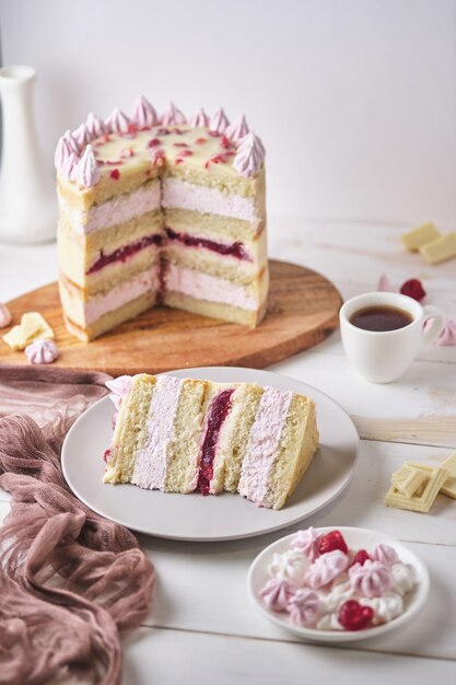 Cake with white chocolate and cherry on a wooden board on a white table. Sliced piece of cake on a plate and a cup of espresso. Dessert decorated with homemade marshmallows.