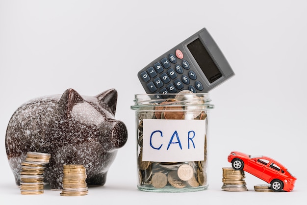 Calculator on glass coins jar with coin stack; car and piggybank against white background