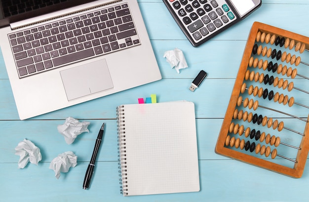 A calculator, a laptop and old abacus lie on a blue wooden table. The crumpled sheets speak of complex calculations. The link between generations.Mockup.