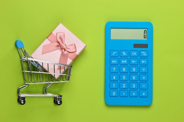 Calculator and shopping trolley with gift box on green. Calculation of the value of the gift.