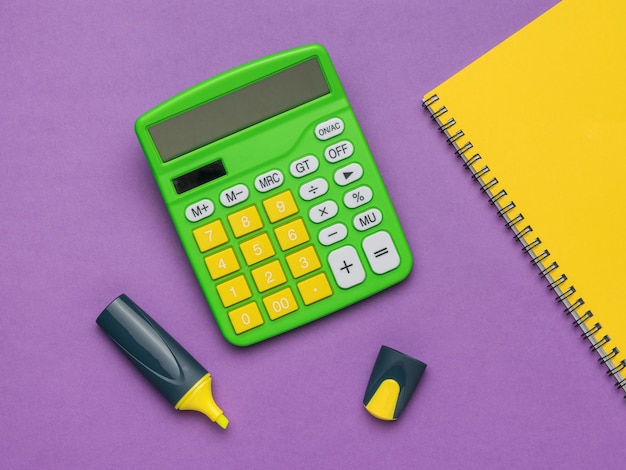 A calculator a yellow marker and a yellow notebook on a purple\
background flat lay