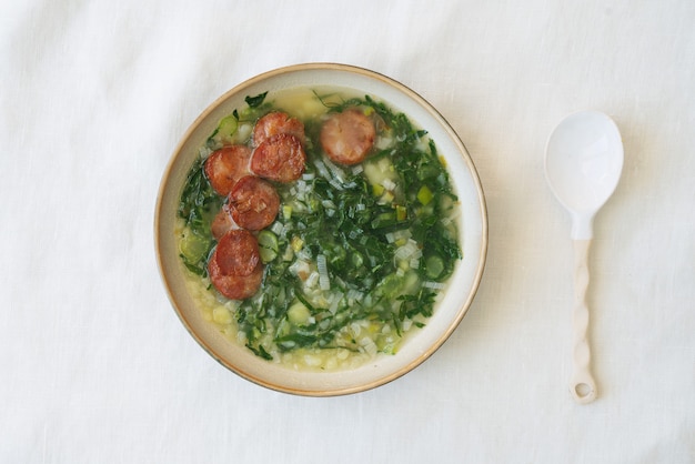 Caldo Verde Soep met groenten en gehakte chorizo bovenop in keramische kom met keramische lepel. Bovenaanzicht