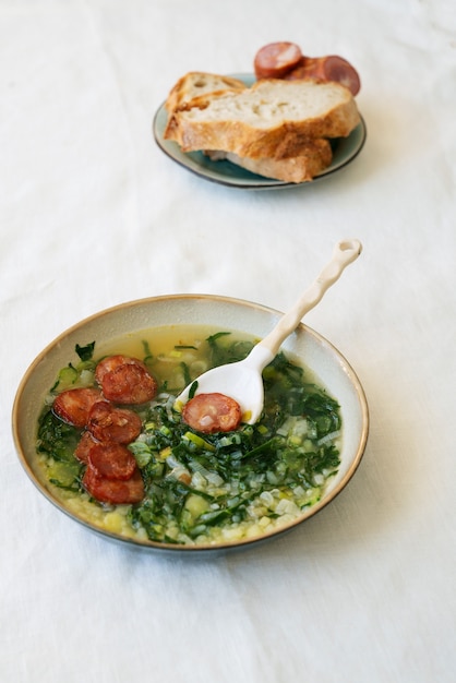 Caldo Verde Soep met groenten en gehakte chorizo bovenop in keramische kom met keramische lepel. Stukjes brood en chorizo op keramische plaat