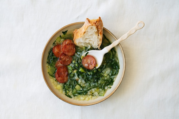 Фото caldo verde суп с зеленью и нарезанным чоризо сверху в керамической миске с керамической ложкой и кусочком хлеба. вид сверху