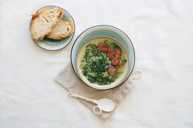 Caldo Verde Суп с зеленью и нарезанным чоризо сверху в керамической миске поверх керамической тарелки на хлопковом мешочке с керамической ложкой. Кусочки хлеба на тарелке. Вид сверху