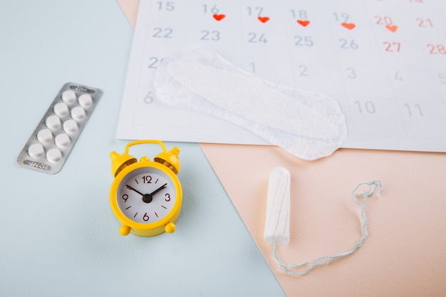 Calendario, tampone di cotone e sveglia gialla su sfondo rosa blu. concetto di ciclo mestruale femminile.