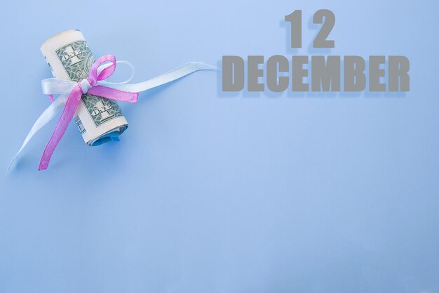 Календарная дата на синем фоне со свернутыми долларовыми купюрами, закрепленными синей и розовой лентой с копией пространства 12 декабря - двенадцатый день месяца
