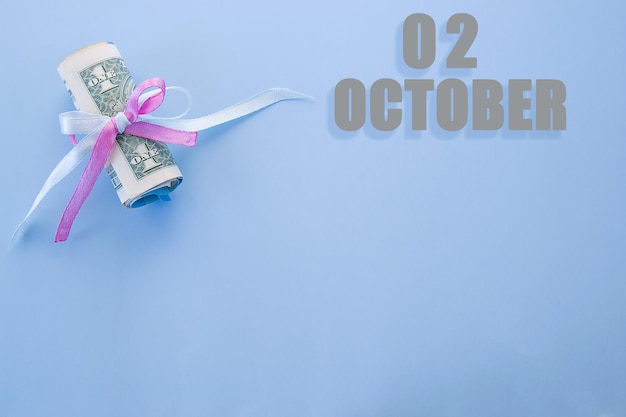 Календарная дата на синем фоне со свернутыми долларовыми купюрами, закрепленными синей и розовой лентой с копией пространства 2 октября - второй день месяца
