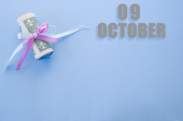 Календарная дата на синем фоне со свернутыми долларовыми купюрами, закрепленными синей и розовой лентой с копией пространства 9 октября - девятый день месяца