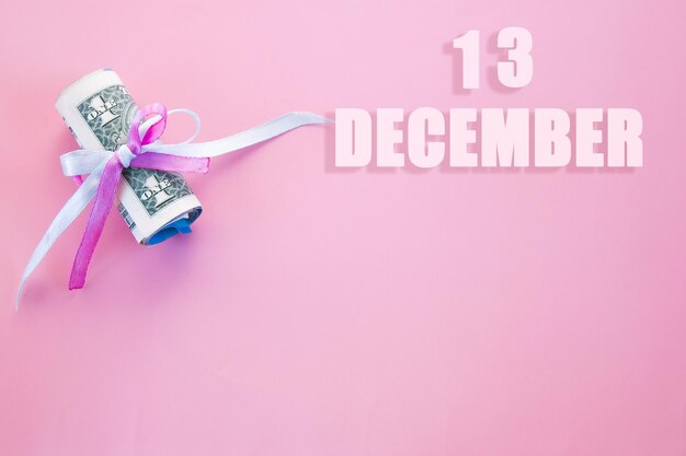 コピースペース付きのピンクとブルーのリボンで固定されたロールアップされたドル札が付いたピンクの背景のカレンダーの日付12月13日は月の13日目です。