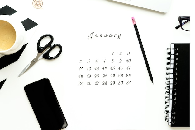 Calendar of January on white desktop flat ley with a cup of coffee and a notebook.