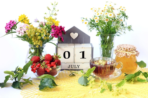 Calendar for July 1 the name of the month of July in English cubes with the numbers 0 and 1 bouquets of wild flowers jam fruit a cup of tea on a yellow openwork napkin