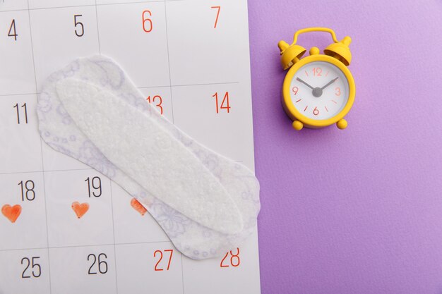 Calendar, menstrual pad and yellow alarm clock on lilac background. Female's menstrual cycle concept.