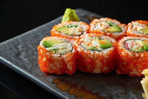 California sushi roll with crab, avocado, cucumber and tobiko caviar served on a plate with wasabi and ginger. Isolation on a black table. Japanese food