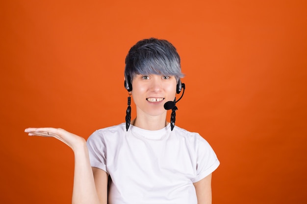 Call center assistant with headphones on orange wall look happy and positive with confident smile holding  hand with empty space on left