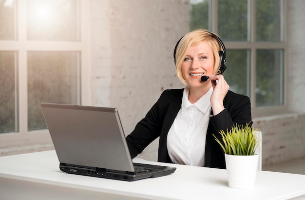 Call center operator blonde vrouw gekleed in zwarte jas en wit overhemd werkt op laptop via headset in wit kantoor