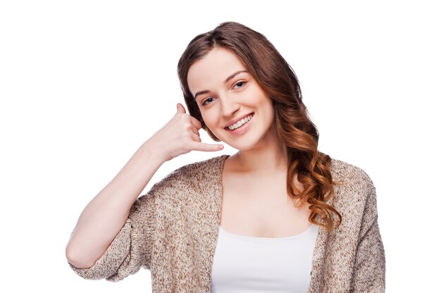 Photo call me! beautiful young smiling woman looking at camera and gesturing mobile phone near ear while standing isolated on white