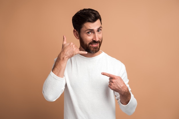 Call me. Portrait of cheerful handsome man standing with telephone hand gesture and smiling to camera, flirting offering to contact by phone. Studio shot isolated on beige background
