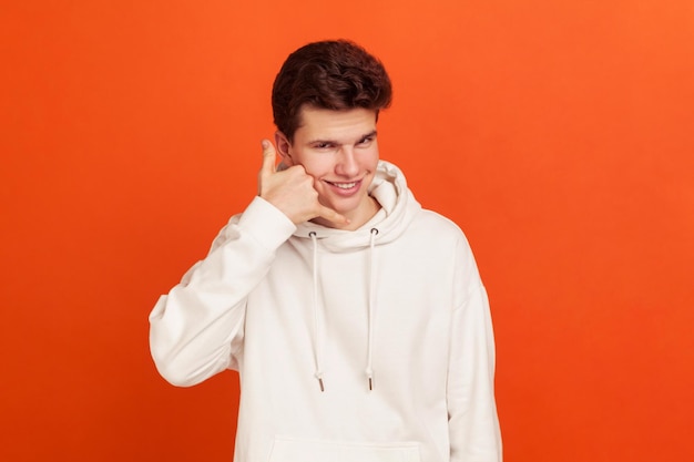 Call me self confident handsome young man in stylish sweatshirt holding fingers near ear highquality mobile communication callcentre indoor studio shot isolated on orange background