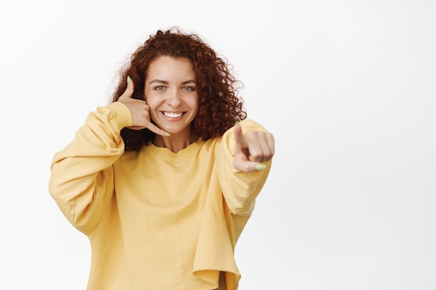 Call me. Smiling young modern woman showing mobile phone hand gesture and pointing, looking happy on white