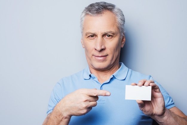 Call this number! Handsome senior man pointing on a business card and smiling while standing against grey background