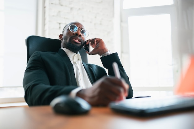 Calling, talking on phone. African-american entrepreneur, businessman working concentrated in office. Looks serios, busy, wearing classic suit. Concept of work, finance, business, success, leadership.