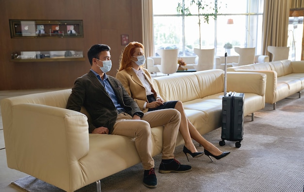 Coppia adulta calma che indossa maschere mediche mentre è seduta su un divano in una hall di un hotel con una custodia per passeggino