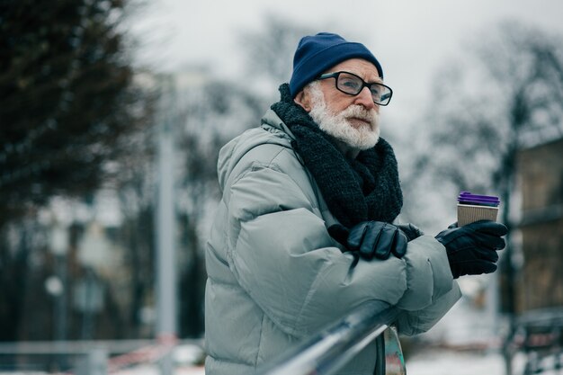 Спокойный пожилой мужчина задумчиво смотрит вдаль, стоя в теплом зимнем пальто с картонной чашкой горячего кофе