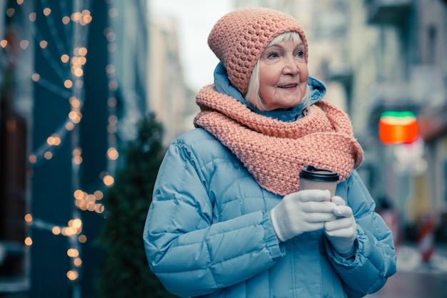 寒い日を歩きながら暖かいコートと一杯のコーヒーを保持している帽子をかぶった穏やかな老婆