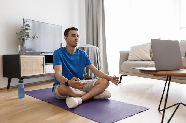 Calm arab man meditating with trainer online via laptop connection using pc in living room sitting in lotus position