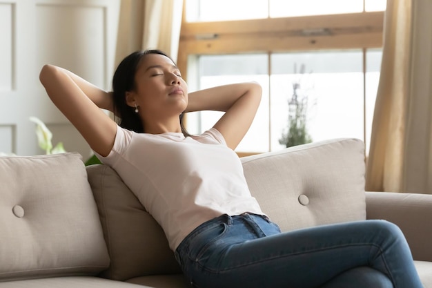 Calm asian young woman sit relax on sofa in living room take\
nap or daydream peaceful vietnamese millennial girl rest on couch\
at home breathe fresh air relaxation stress free concept