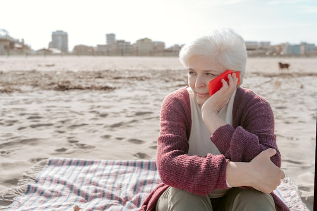 Calm attractive senior woman sitting alone on the sand beach and calling on the smartphone