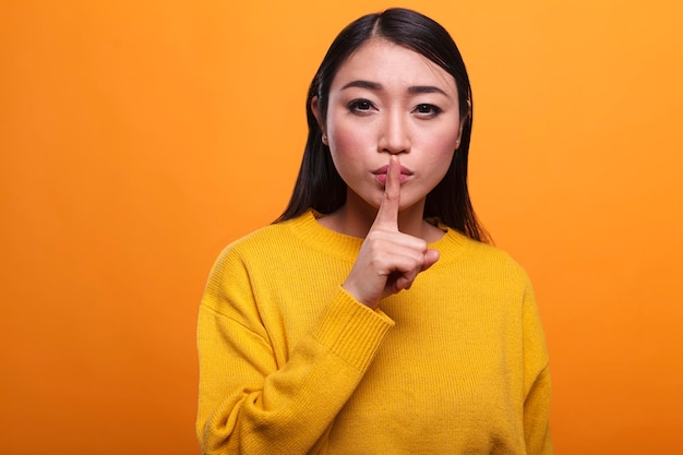 Calm attractive women wearing yellow sweater making hush
gesture with forefinger on orange background. mysterious person
touching lips with index finger indicating silence and
secrecy.