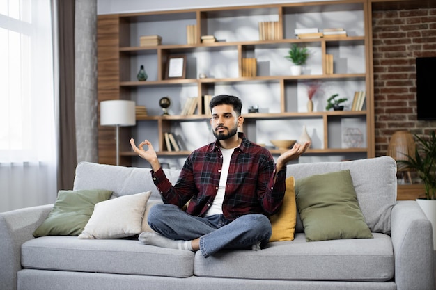 Calm of bearded young man doing yoga lotus pose to meditation and relax on couch during work