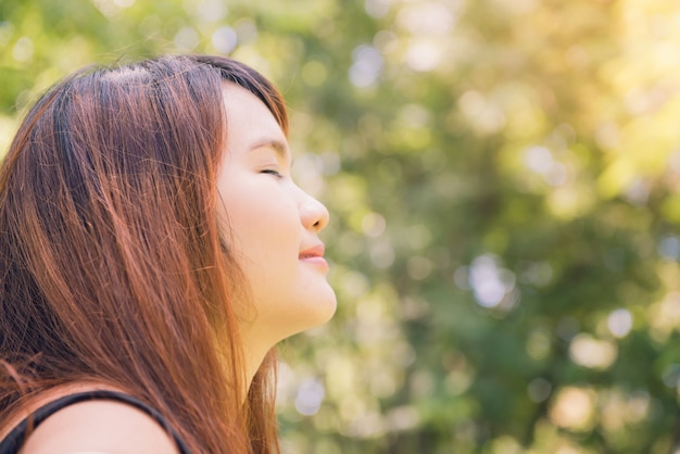 Calm beautiful smiling young woman with ponytail enjoying fresh air outdoor, relaxing with eyes closed, feeling alive, breathing, dreaming. copy space, green park nature background. side view portrait