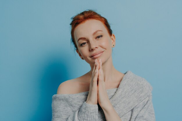 Calm beautiful young woman with red hair in bun making wish with palms pressed asking for good luck