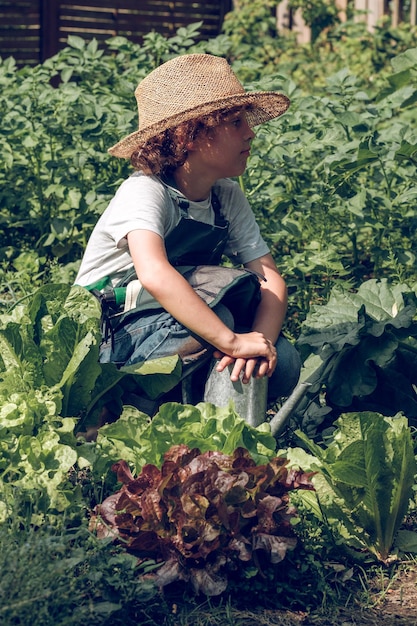 Calm boy in hat squatting on ground with metal watering can near plants while looking away in garden