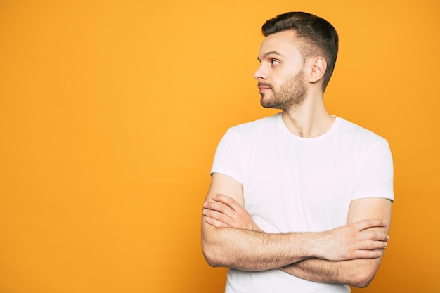 A calm boy with a serious face is standing in front of spicy-orange background in white T-shirt and his hands crossed on his breast