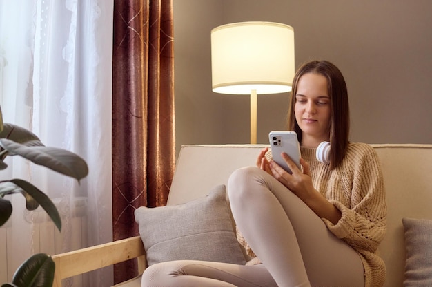 Calm brown haired young Caucasian woman using mobile phone while sitting couch at home scrolling online browsing web pages checking social network or texting with friends