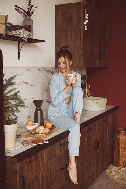 Calm brunette lady holding a cup of tea while sitting one tabletop in the kitchen.